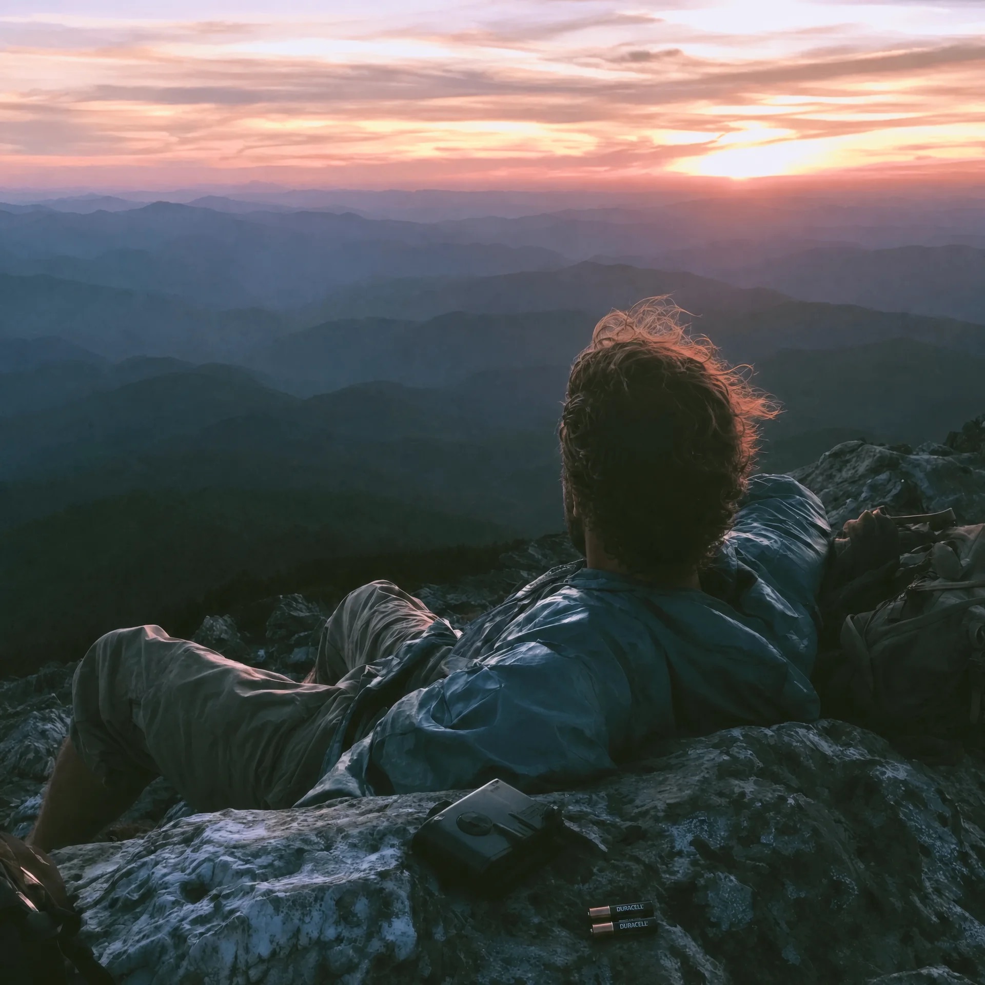 A hiker resting on a summit at sunset, a Duracell battery beside her
