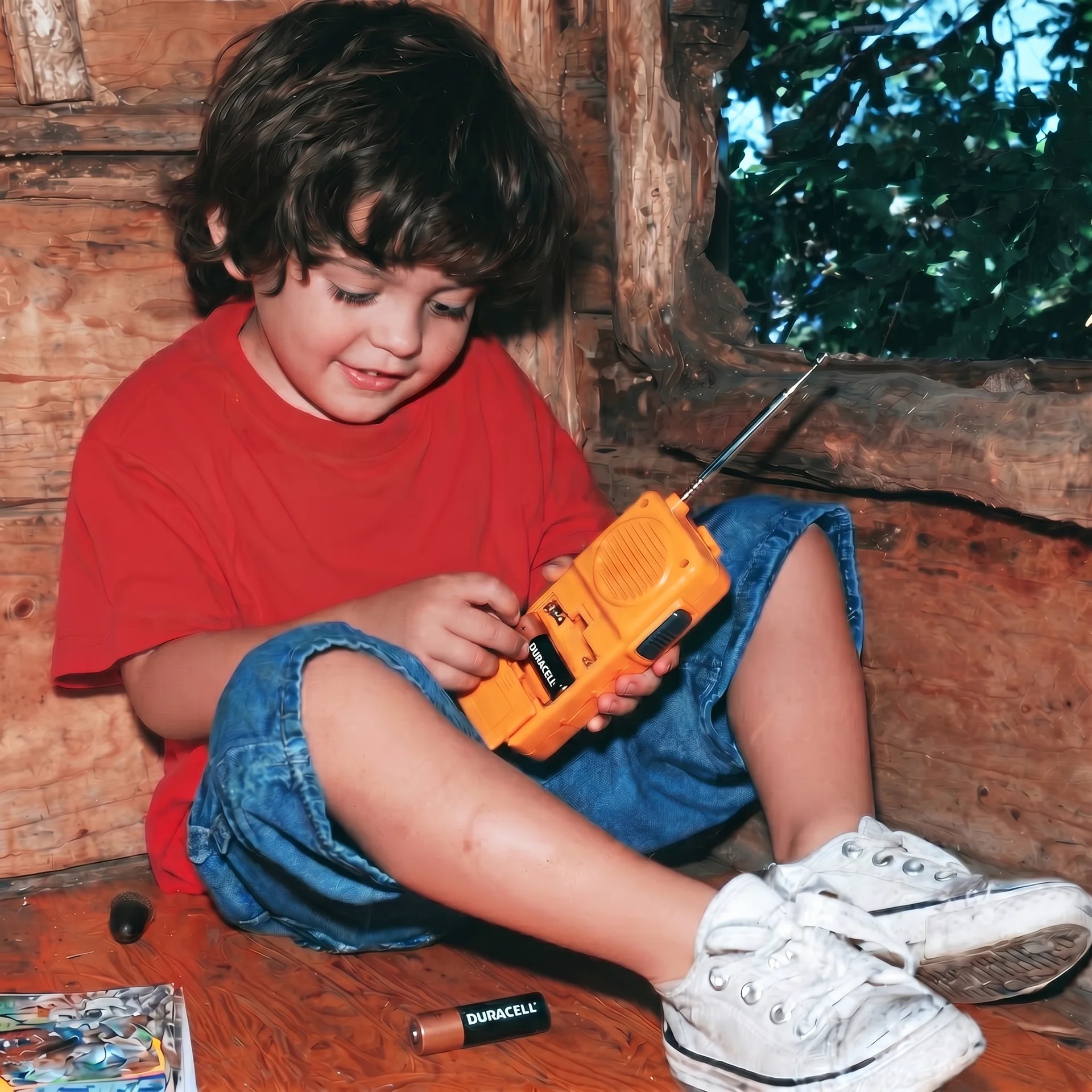 A boy in a red shirt playing with an orange walkie-talkie, a Duracell battery at his feet