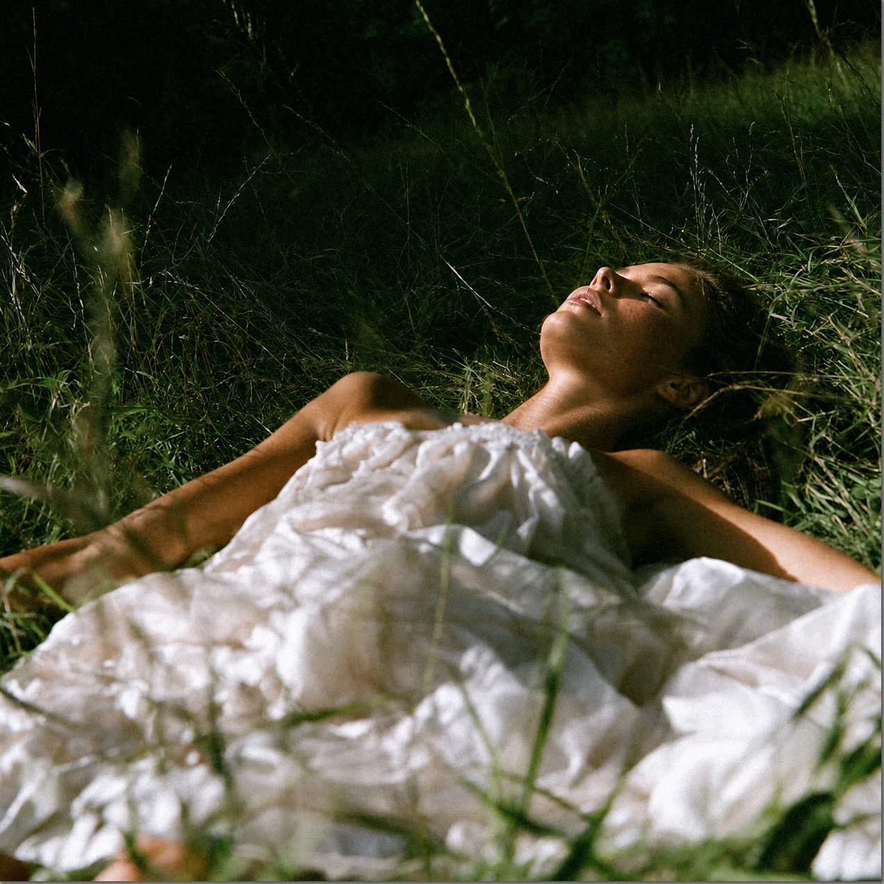 A woman lying in tall grass on white linen, face turned to the sky