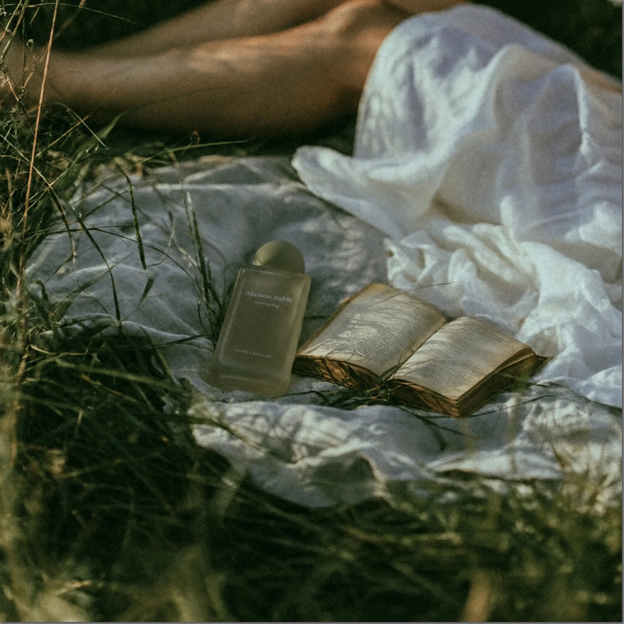 A fragrance bottle and an open book on white linen in the grass