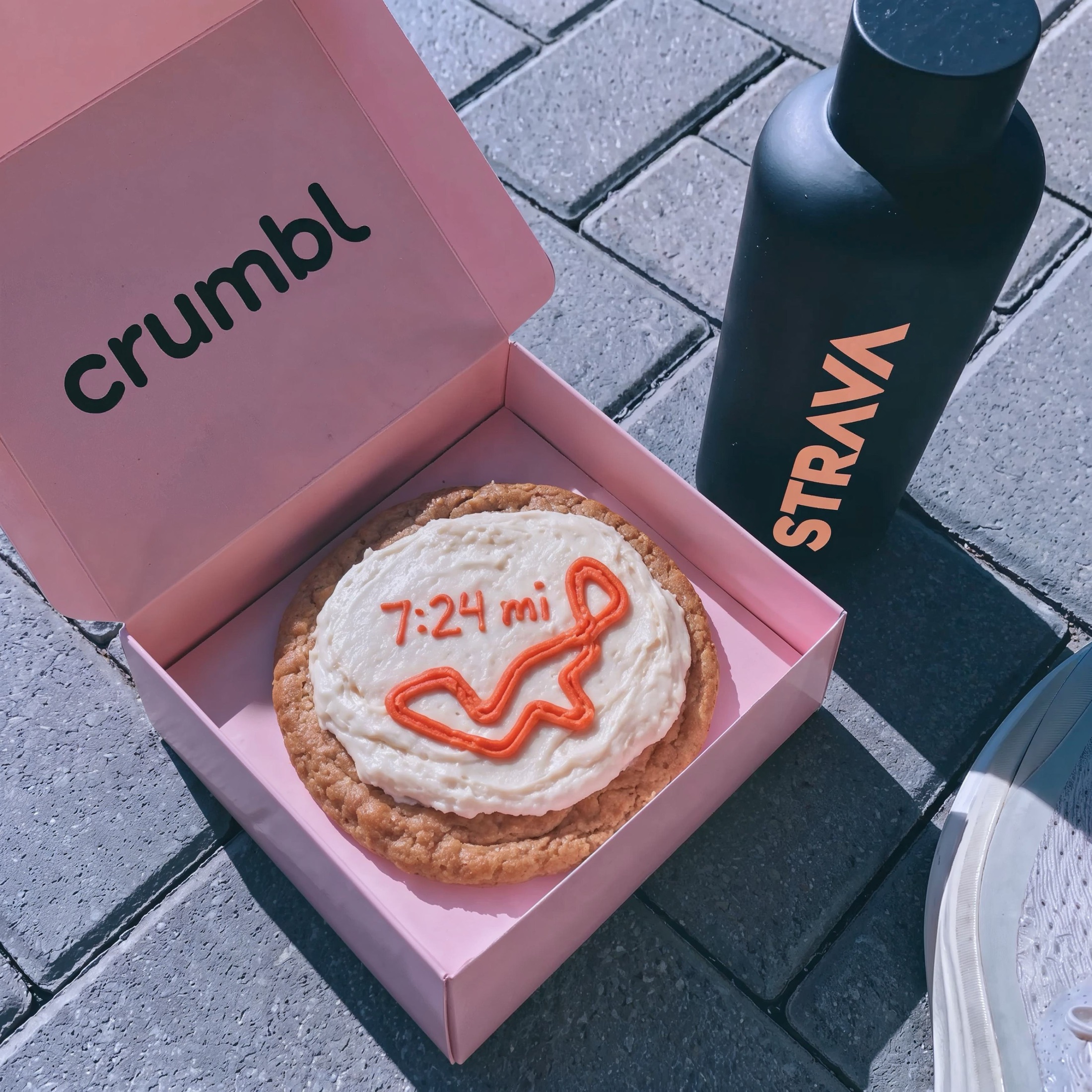 A pink Crumbl box holding a cookie iced with a running route, beside a Strava bottle
